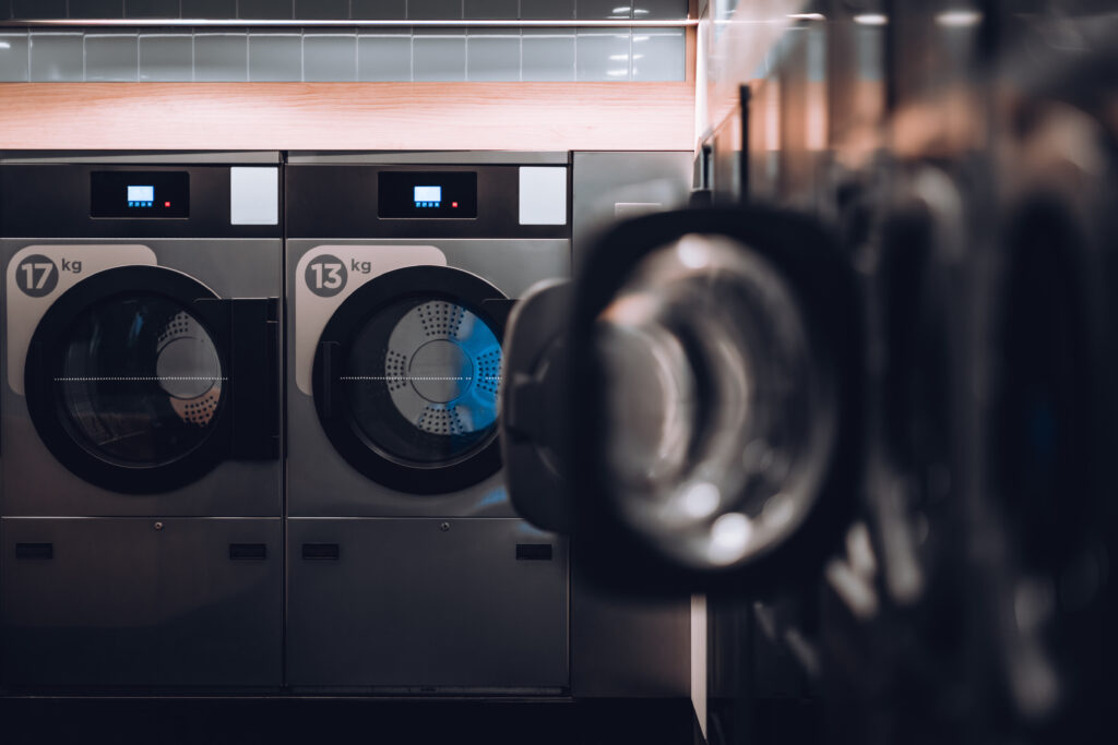 A dark modern room of a publiс laundry with perpendicular rows of washing machines with some of the doors opened and tumble dryers, selective focus on the far wall, shallow depth of field
